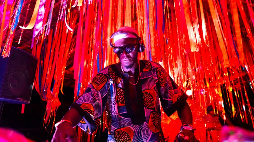 Porträt DJ Emo Rugene für die Lange Nacht der Museen 2026 – Person mit Kopfhörern und Sonnenbrille in bunter Kleidung, umgeben von roten und orangenen Bändern, spielt Musik.