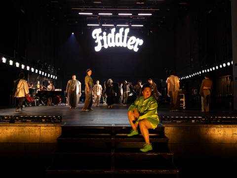  – Bühne mit mehreren darstellenden Personen, Person in grünem Oberteil sitzt vorne, Leuchtschrift "Fiddler" im Hintergrund.