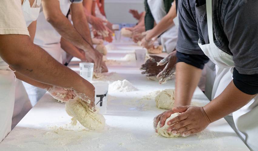 Tiziano Cruz_Bread Workshop – Mehrere Menschen kneten Teig auf einem langen Tisch. Alle tragen Schürzen. Im Hintergrund stehen Becher mit unleserlichem Text.