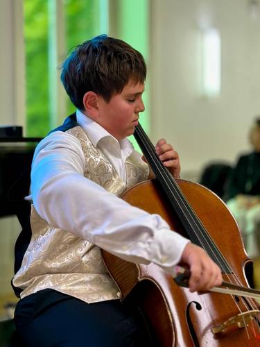 Kind spielt Cello, trägt eine gemusterte Weste über einem weißen Hemd. Im Hintergrund unscharfe grüne und weiße Flächen.