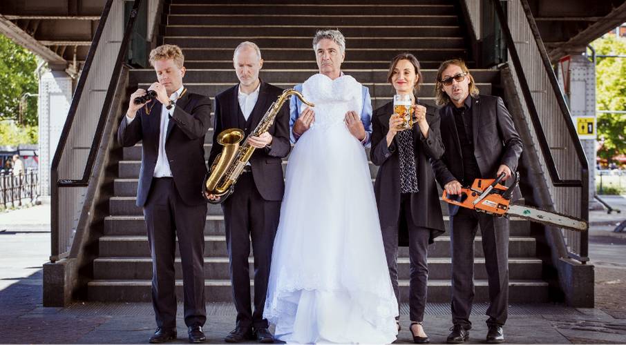 Fünf Personen auf einer Treppe, eine Person in einem Hochzeitskleid, die anderen elegant gekleidet mit Kamera, Saxophon, Bier, Kettensäge.