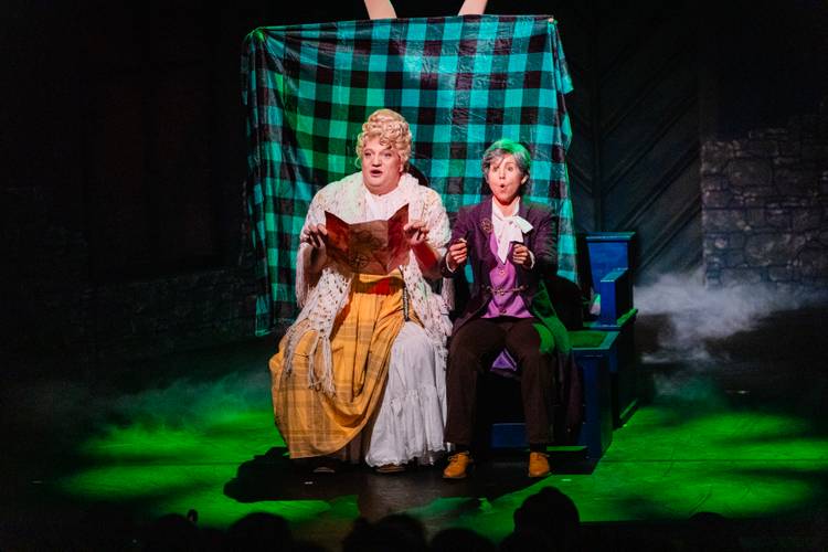 Dracula – A Comedy of Terrors – Zwei Darsteller*innen auf einer Bühne vor kariertem Vorhang, eine Person mit langem Rock und Spitze, die andere in Anzug mit Krawatte, beide mit erstaunten