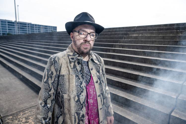 Person mit Brille, Hut und Schlangenlederjacke vor Treppe im Freien; Rauch im Vordergrund.