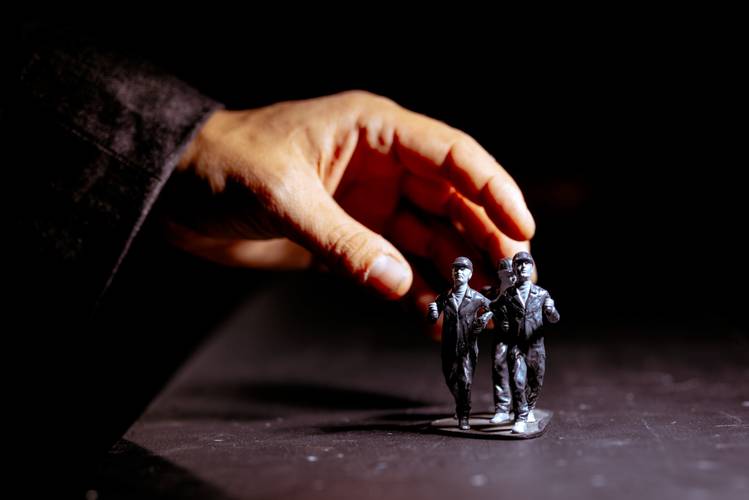 Transport: Tipping Point – Eine Hand greift nach zwei kleinen, grauen Spielfiguren von Arbeiter*innen in Uniformen auf einem dunklen Hintergrund.