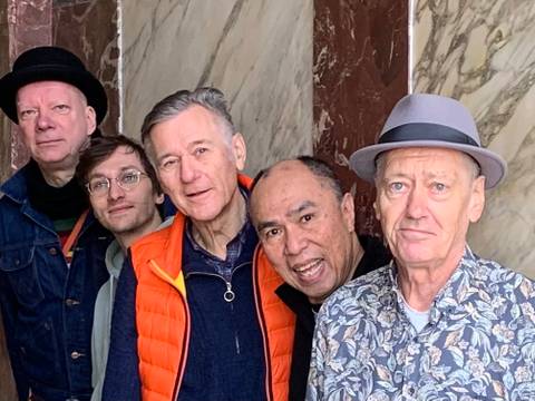 George Kranz & friends – Fünf Personen stehen nebeneinander, manche tragen Hüte und Brillen. Eine Person trägt eine orange Weste, eine andere ein Hemd mit Blumenmuster. Marmorsäulen im