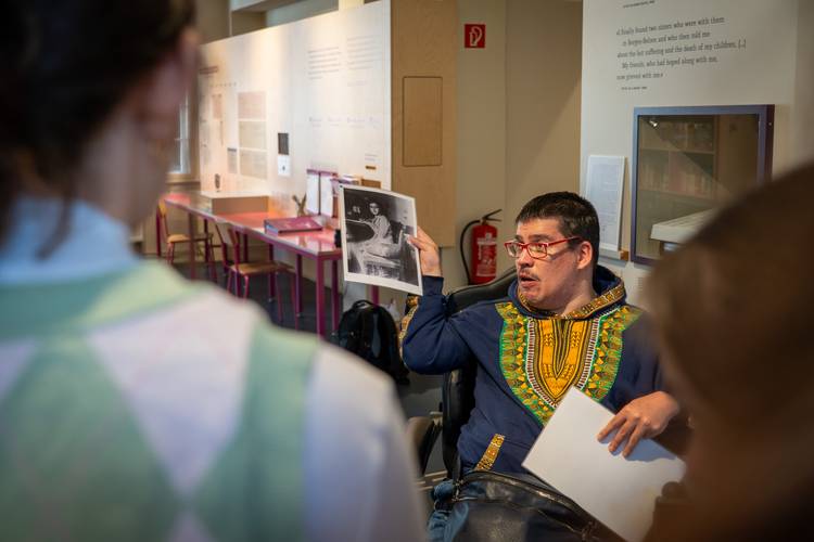 Peer Guides begleiten durch die Ausstellung – Mann in Ausstellung zeigt einer Gruppe ein historisches schwarz-weiß-Foto von Anne Frank.