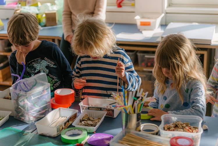 Drei Kinder basteln an einem Tisch mit bunten Bastelmaterialien wie Pfeifenreinigern, Holzchips, Klebeband und Stiften in einem hellen Raum.