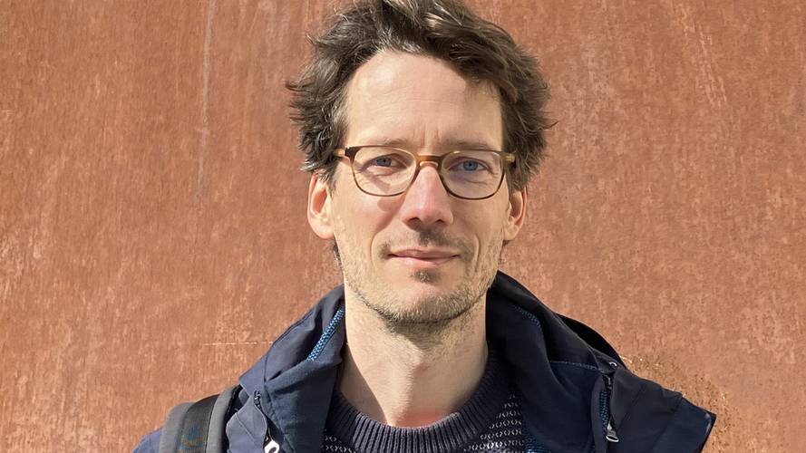 Christian Staas – Person mit Brille, dunkler Jacke und Pullover mit Krausmuster vor rostfarbener Metallwand. Sie hat braune Haare und traegt einen Rucksack.