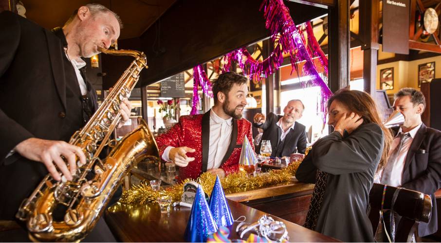 Person in rotem Paillettenjackett lacht an einer Bar, spielt Saxophon; weitere Personen feiern mit Partyhueten und Dekorationen, Geraeuschempfindlichkeit