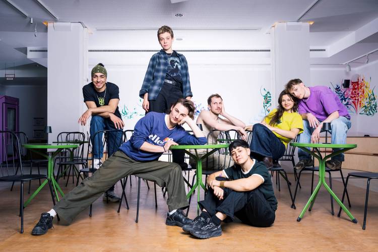 Sieben Personen posieren lässig auf und um grüne Tische und schwarze Stühle, bunte Wandmalerei im Hintergrund. Eine Person trägt ein Shirt mit "ellesse".