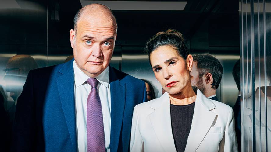 Zwei Personen in Anzug und Blazer stehen nebeneinander in einem Aufzug, der metallene Wände hat.
