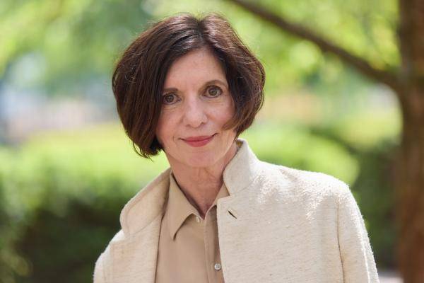Prof. Dr. h.c. Jutta Allmendinger – Eine Person mit schulterlangem, glattem Haar, hellem Mantel und beigem Hemd steht draußen vor unscharfem, grünem Hintergrund.