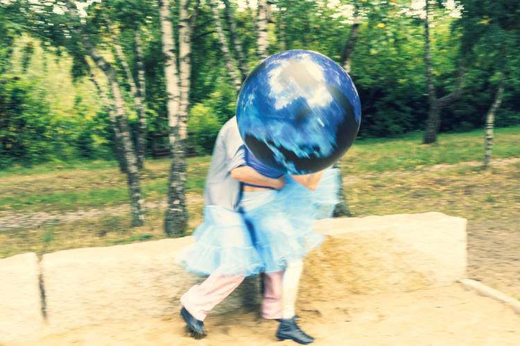 Person trägt ein blaues Tutu und hält einen großen aufblasbaren Globus. Hintergrund mit Bäumen und Sandfläche.