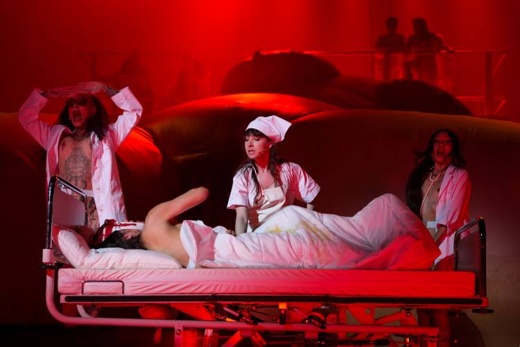 Person in Krankenhausbett, umgeben von drei Darsteller*innen in weißer Kleidung. Roter Hintergrund, dramatische Beleuchtung.