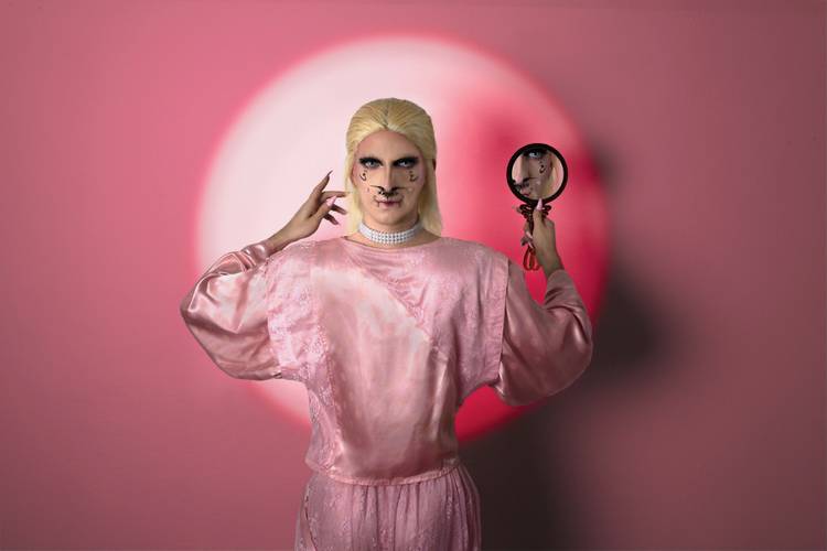 Eine Person in einem rosa Satinoutfit und blonder Perücke steht vor rosafarbenem Hintergrund mit kreisförmigem Lichtschein und hält einen kleinen Spiegel;