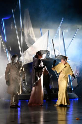 Don Giovanni – Drei Personen in langen Mänteln und Netzhüten stehen auf einer Bühne mit futuristischer Kulisse aus leuchtenden Stangen und weißen Tüchern.