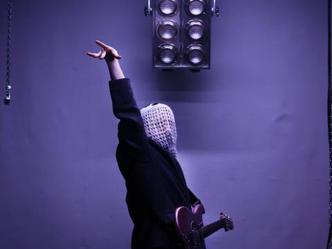  – Eine Person mit gehäkeltem Tuch über dem Gesicht hält eine E-Gitarre und zeigt mit dem erhobenen Arm nach oben. Überhead ist eine große Lampe.