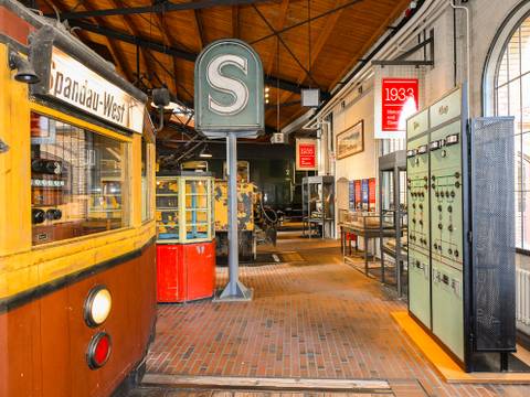 Der Führerstand eines Berliner S-Bahn-Triebwagens von 1927 erinnert an die Einführung der Stadt-, Ring- und Vorortbahnen nach dem ersten Weltkrieg. Sie werden bis heute über eine seitliche Stromschiene versorgt, die speziell hierfür entwickelt wurde – Der rot-gelbe Führerstand eines S-Bahnwagens steht im Vordergrund, dahinter steht ein großes S-Bahn Schild in der Ausstellung.