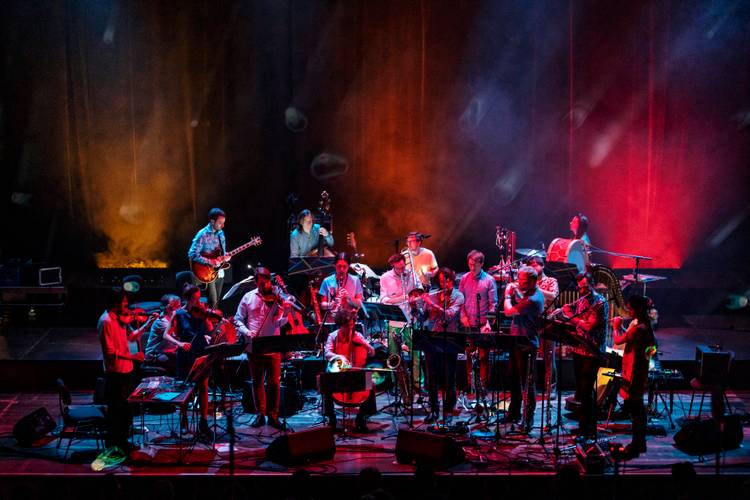 AMEO Kosmostage, Foto: Gianmarco Bresadola – Große Musikgruppe auf einer Bühne, spielt verschiedene Instrumente wie Gitarre, Geige, Trompete und Schlagzeug; bunte Beleuchtung in Rot und Blau.