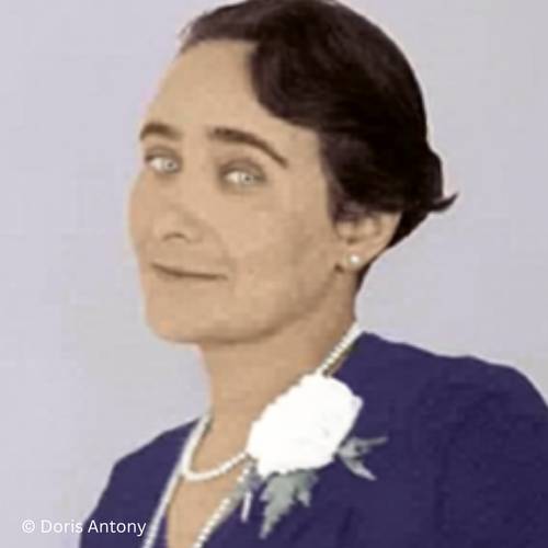 Gabriele Tergit – Person mittleren Alters mit kurzem dunklem Haar, Perlenkette und Ohrringen, lila Kleid mit weißer Blume. Text: © Doris Antony.