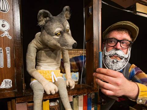  – Wolfspuppe in gelben Shorts sitzt auf einem Regal mit Buechern. Person mit Brille und Papierbart haelt das Regal.