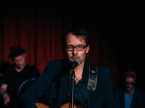 Jan Böttcher – Person mit Brille und Anzug spielt Gitarre und singt in ein Mikrofon vor rotem Vorhang; zwei Personen im Hintergrund.