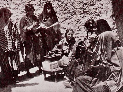 Foto aus Brigitte Schiffers Dissertation "Die Oase Siwa und ihre Musik" (1936) – Sechs verschleierte Personen stehen und sitzen um eine Kamera auf einem Tisch vor einer Steinwand.