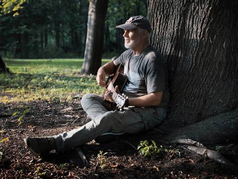  – Person mit grauem Bart, dunklem T-Shirt und Cap sitzt im Wald mit Gitarre an einen Baum gelehnt. Sonnenlicht fällt durch das Blätterdach.
