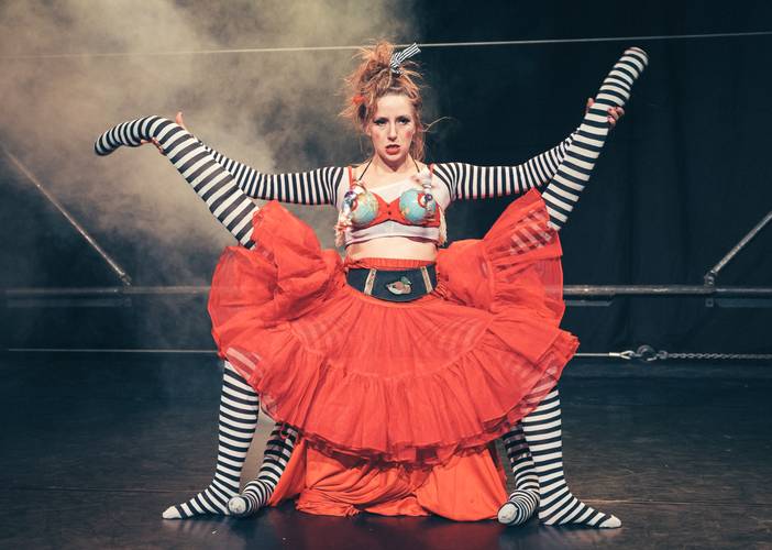 Barbaren Barbies: A wild women circus – Person mit rotem Rock und gestreiftem Oberteil sitzt auf schwarz-weiß gestreiften Beinen, rauchiger Bühnenhintergrund.