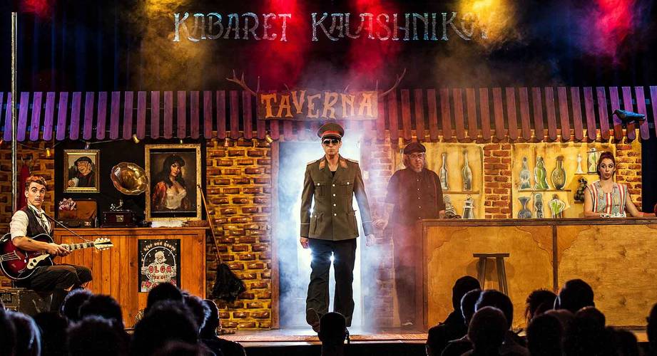 Kabaret Kalashnikov – Bühne mit drei Personen vor einer gemauerten Kulisse. In der Mitte eine Person in Militäruniform, links ein Gitarrist, rechts eine Person hinter einer Bar.