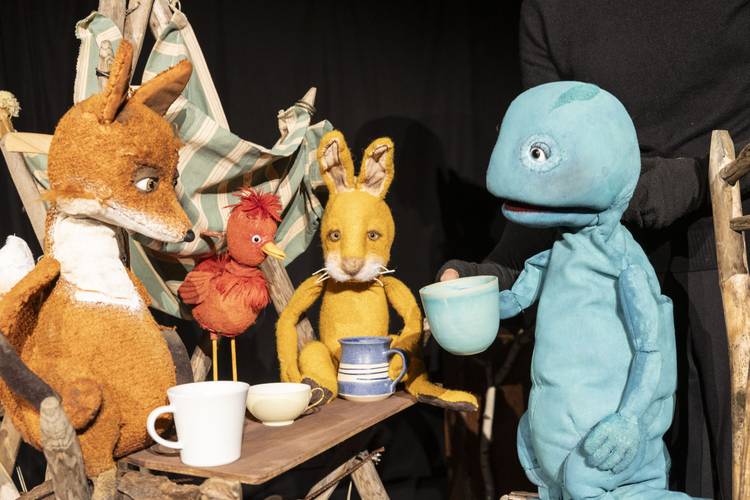Der Koffer – Vier bunte Puppen: Fuchs, Vogel, Hase, blaues Wesen, sitzen um Tisch mit Tassen. Eine person hält das blaue Wesen.