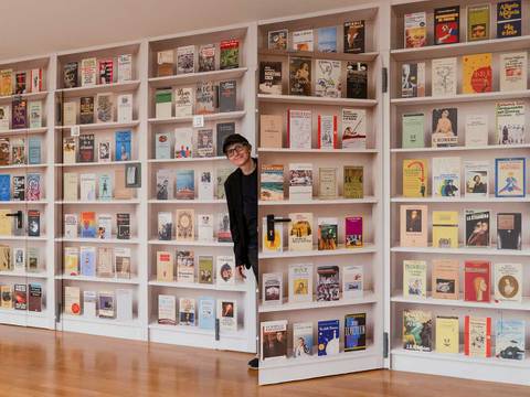 Daniela Comani, Orlando's Library, Wandtapete, 254 x 700 cm, MAMbo – Museo d’Arte Moderna, Bologna, seit 2023 – Person blickt zwischen Buecherregalen hervor; Regale voller bunter Buecher in weißem Raum. Holzboden. Einige Buchcover beschriftet, z. B. "Chekhov".