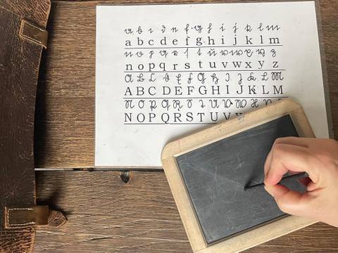 Um die Sütterlin-Schrift zu lernen, schrieben Kinder früher mit einem Griffel auf einer Schiefertafel. – Eine Person schreibt mit einem Griffel auf einer Schiefertafel