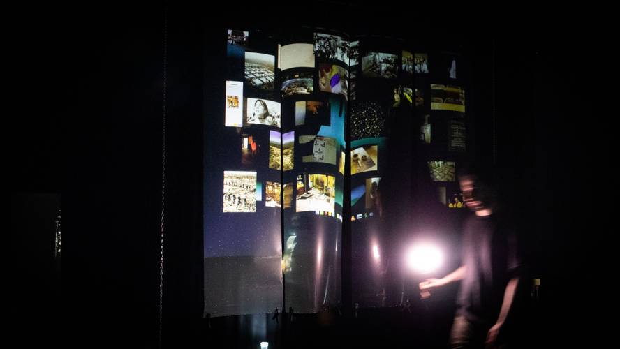 Mei Liu, Homesick for Another World (2025). Foto: Jia Y. Ding – Beleuchtete Projektionsflaeche mit vielen Fotos in Reihen. Unscharfe Person rechts haelt eine Lampe. Umgebung ist dunkel.