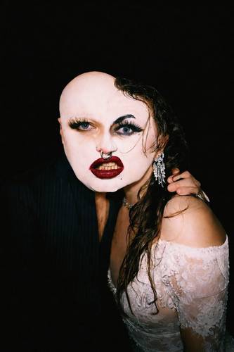 Erratum – Porträt einer Person mit weiß geschminktem Gesicht, tiefroten Lippen, Nasenring und dramatischen Wimpern vor schwarzem Hintergrund; eine Gesichtshälfte ist kahl, die andere von nassem dunklem Haar bedeckt, dazu ein weißes Spitzenkleid und funkelnder Ohrring.