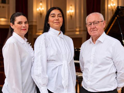  – Drei Personen in weißen Hemden stehen in einem eleganten Raum mit Kronleuchtern, ein Flügel im Hintergrund.
