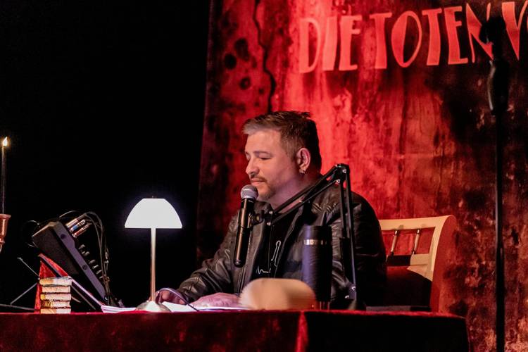 Person mit Mikrofon und Kopfhörern an einem Tisch, beleuchtet von Lampe, im Hintergrund ist der Schriftzug "DIE TOTEN V..." sichtbar.