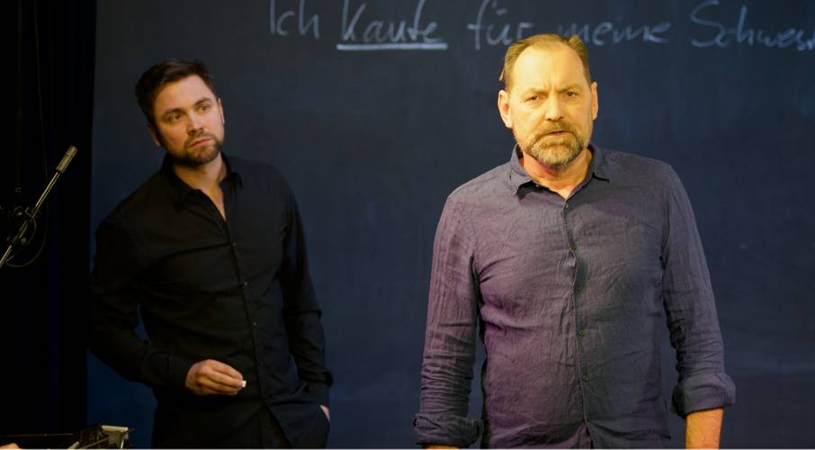 Zwei männlich gelesene Personen vor einer Tafel mit der Aufschrift "Ich kaufe für meine Schwes…".