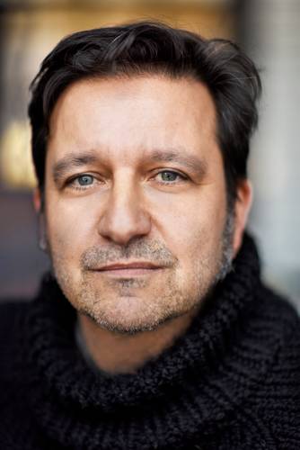 Julian Voloj – Person mit dunklen Haaren und Dreitagebart, trägt einen schwarzen Strickpullover, schaut ernst. Hintergrund unscharf.