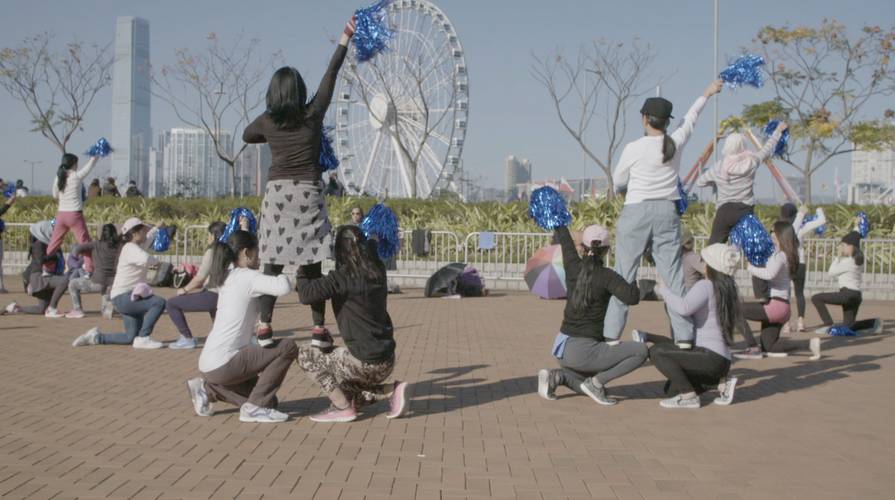 Stephanie Comilang, Come to Me, Paradise, 2017 – Video-Still: Eine Gruppe von Menschen im Freien mit blauen Pompons, einige kniend und andere stehend, vor einem Riesenrad und Hochhäusern in einer Stadtlandschaft.