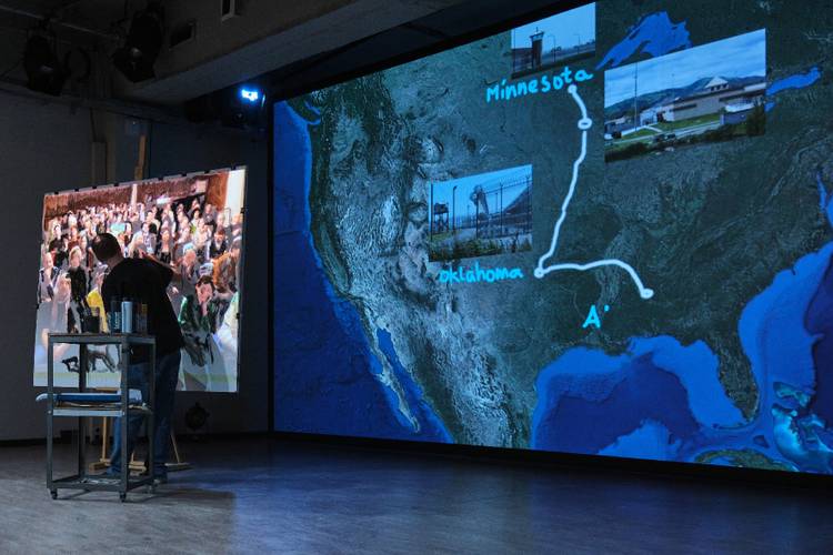 Person malt vor großem Bildschirm; USA-Karte mit Fotos: Minnesota, Oklahoma. Weiße Route verbindet Orte. Publikum auf Leinwand links.