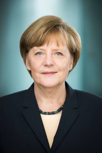 Angela Merkel – Eine weiblich gelesene Person mit kurzem Haar, in dunklem Blazer und Halskette, schaut direkt in die Kamera. Hintergrund unscharf und blau-grau.