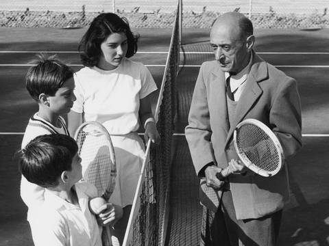 Moderne unter kalifornischer Sonne: Arnold Schönberg mit seinen Kindern Lawrence, Ronald und Nuria beim Tennis in Santa Monica, 1948 – Mann im Anzug mit Tennisschläger spricht über ein Tennisnetz hinweg mit drei Kindern in Tennisbekleidung