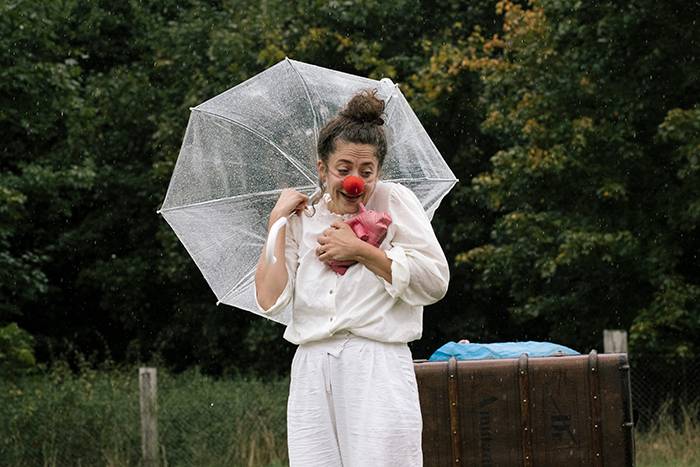 Person mit roter Clownsnase hält durchsichtigen Regenschirm und roten Stoff, trägt weiße Kleidung. Hintergrund aus Bäumen und einer Holzkiste.