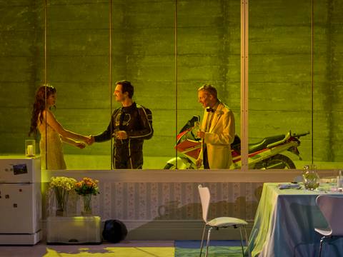 Die Glasmenagerie – Eine Frau im Pünktchenkleid und ein Motorradfahrer geben einander lächelnd die Hand, während ein Mann im Anzug zusieht