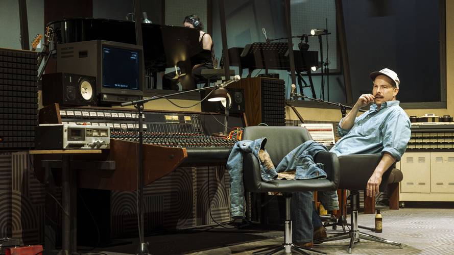 Stereophonic – Tonstudio mit Mischpult und Monitor. Person mit Kappe sitzt lässig auf einem Stuhl. Im Hintergrund spielt eine Person Klavier.