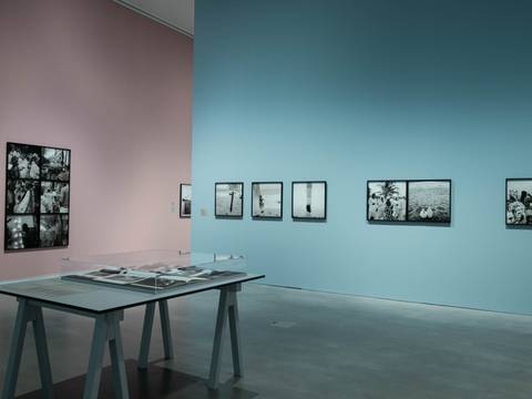 Ausstellungsansicht „Akinbode Akinbiyi. Being, Seeing, Wandering“ – In einer gläsernen Auslage in der Mitte des Ausstellungsraums liegen Fotobücher. Im Hintergrund sind verschiedene Schwarz-Weiß-Fotografien an den pastel-rosa und pastel-blauen Ausstellungswänden zu sehen.