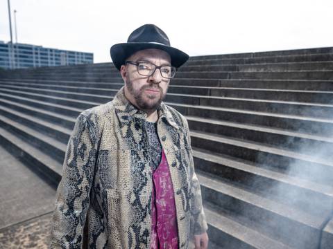  – Person mit Brille, Hut und Schlangenlederjacke vor Treppe im Freien; Rauch im Vordergrund.