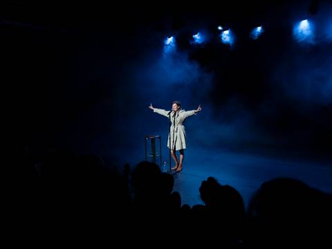  – Eine Person in einem Bühnenraum steht sprechend am Mikrofon mit erhobenen Armen, während blaues Bühnenlicht auf sie fällt. Ein Hocker steht daneben.