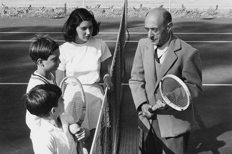 Moderne unter kalifornischer Sonne: Arnold Schönberg mit seinen Kindern Lawrence, Ronald und Nuria beim Tennis in Santa Monica, 1948 – Mann im Anzug mit Tennisschläger spricht über ein Tennisnetz hinweg mit drei Kindern in Tennisbekleidung